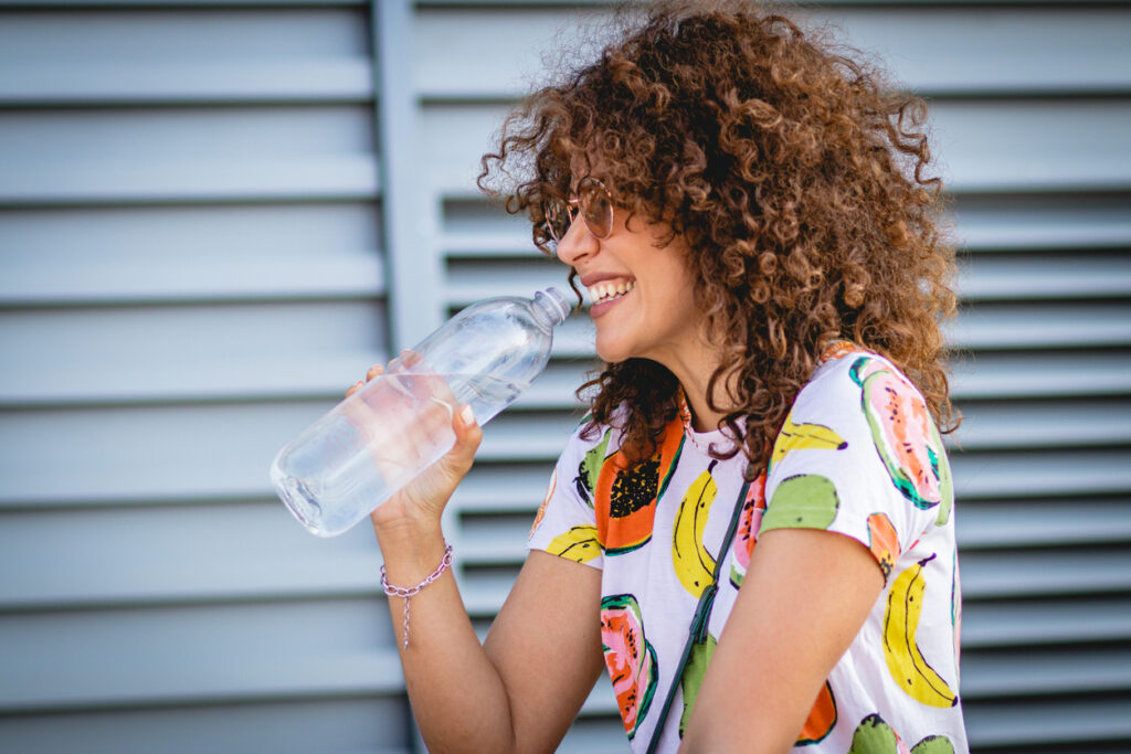 Curly-haired woman laughing while holding a bottle of water in the summer.