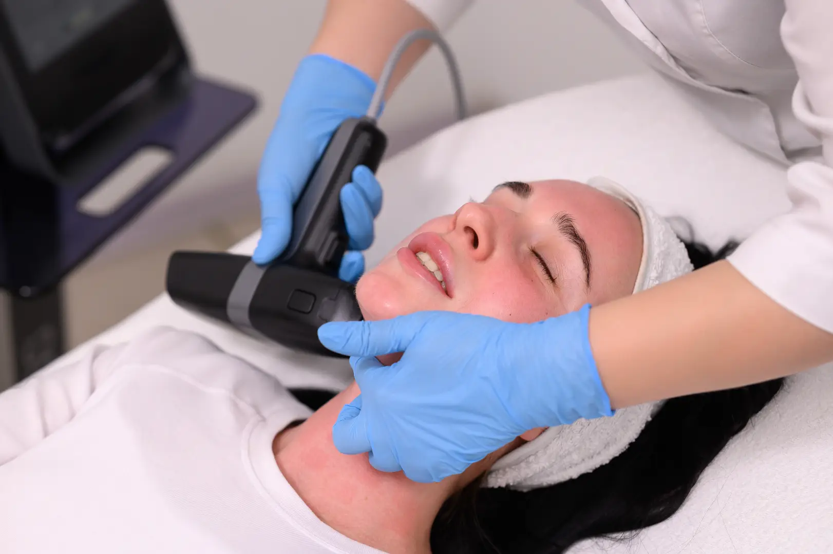 Blue-gloved practitioner presses a black handheld device against a reclining woman’s jawline.